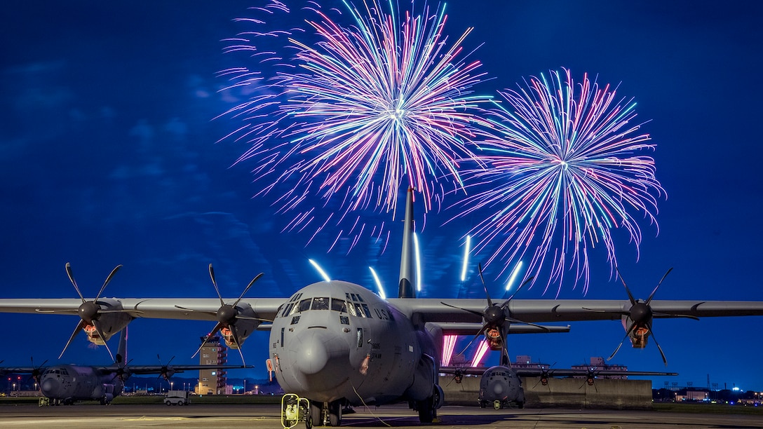 Fireworks go off behind a C-130J Super Hercules aircraft sitting on a flightline.