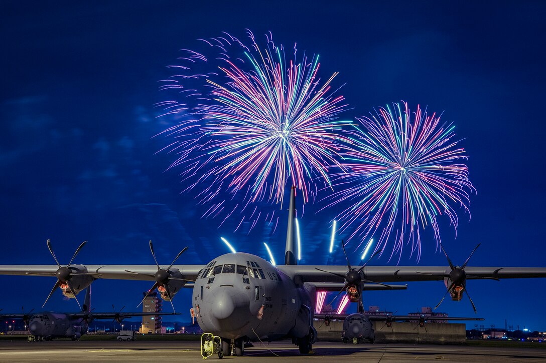 Fireworks go off behind a C-130J Super Hercules aircraft sitting on a flightline.