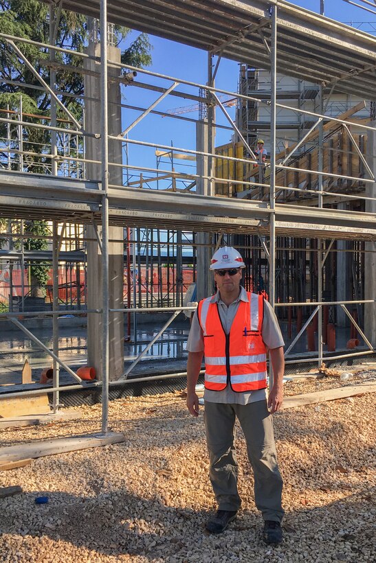 A man stands in front of a construction site, wearing a hard hat and safety vest.