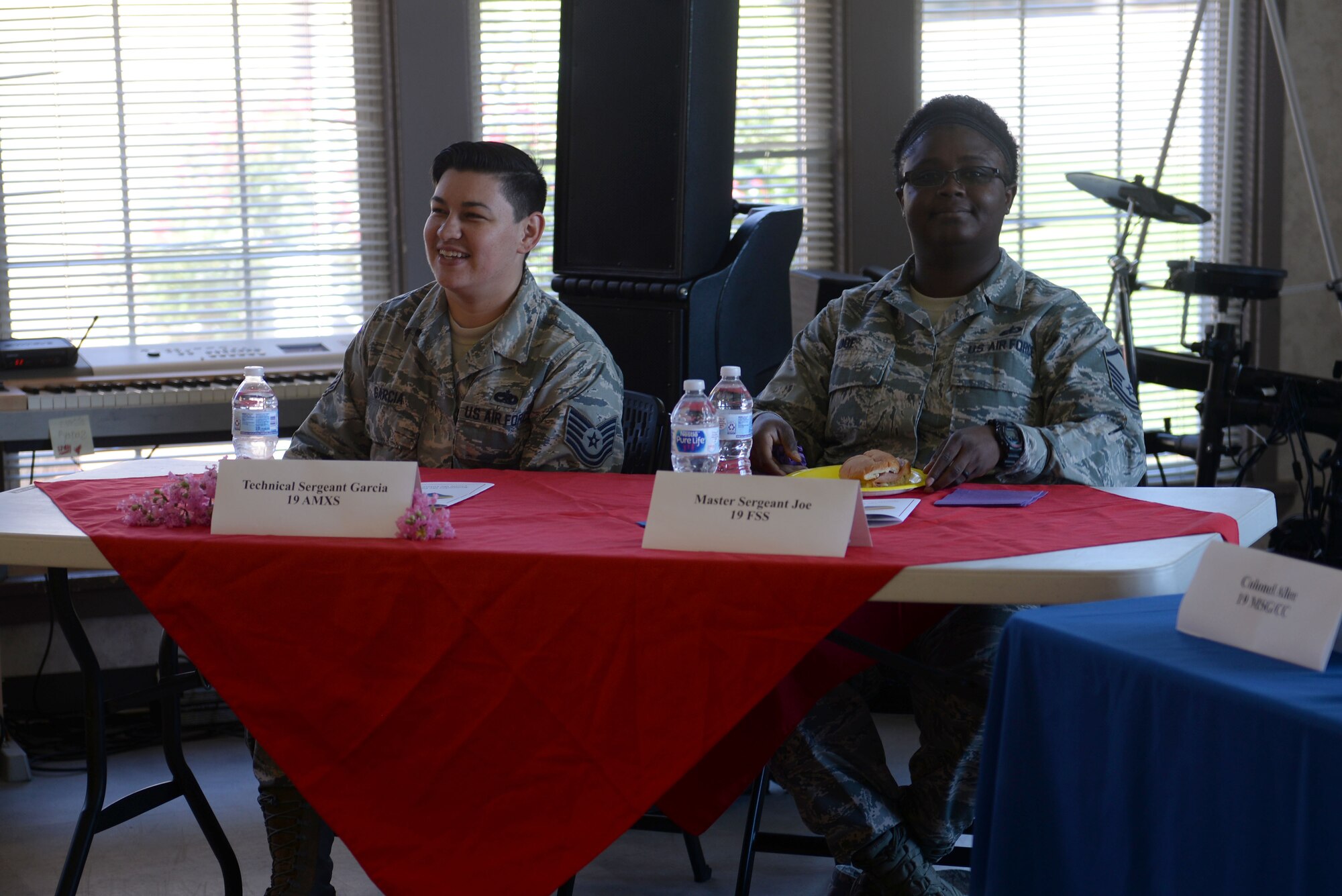 Two women wearing the Airmen Battle Uniform sit at a table with a red table cloth and smile at the camera.