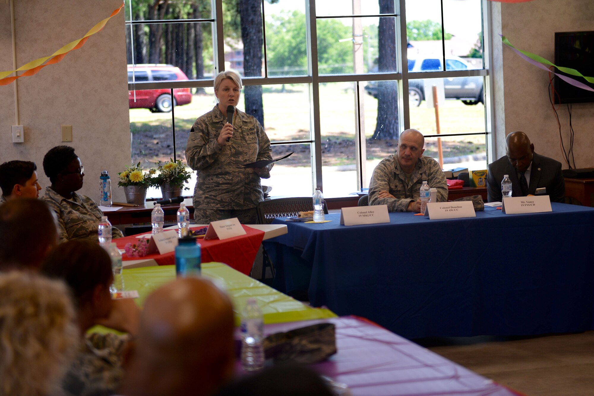 A woman wearing the Airman Battle Uniform speaks into a microphone into a circle of people also wearing the Airman Battle Uniform. They are sitting around tables with table cloths that are each a different color of the rainbow.
