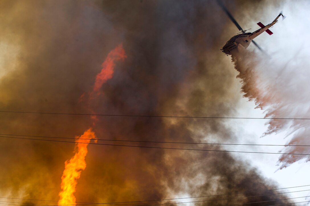 A Marine Corps UH-1 Huey helicopter drops water from a bucket firefighting system onto a wildfire.