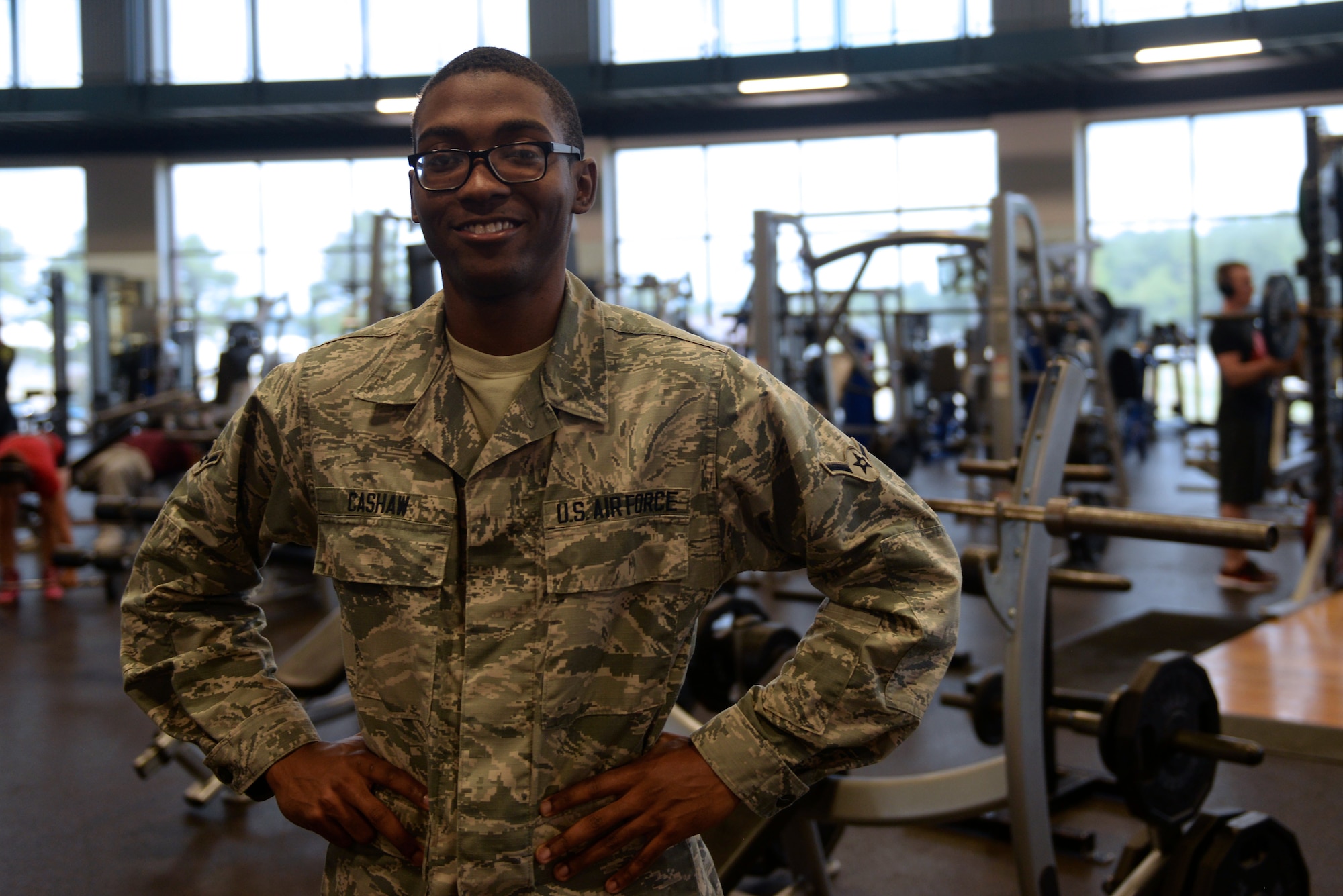 A man wearing the Airman Battle Uniform has his hands on his hips while looking at the camera with exercise equipment behind him. He's wearing glasses.