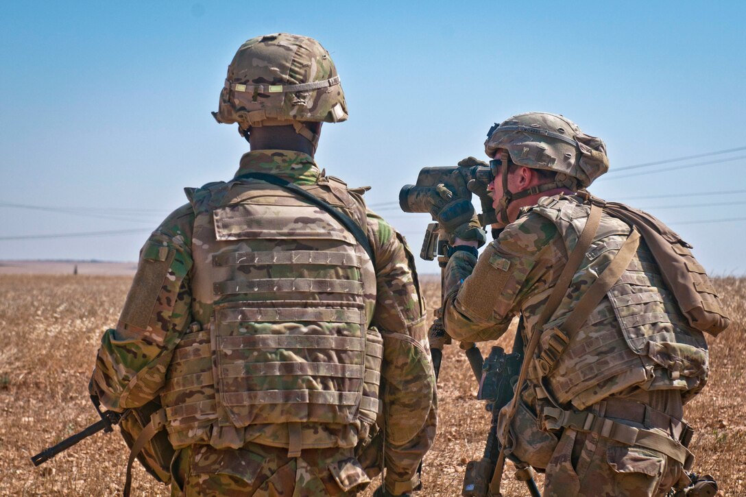 A U.S. soldier looks through a scope at the Turkish soldiers conducting a patrol.