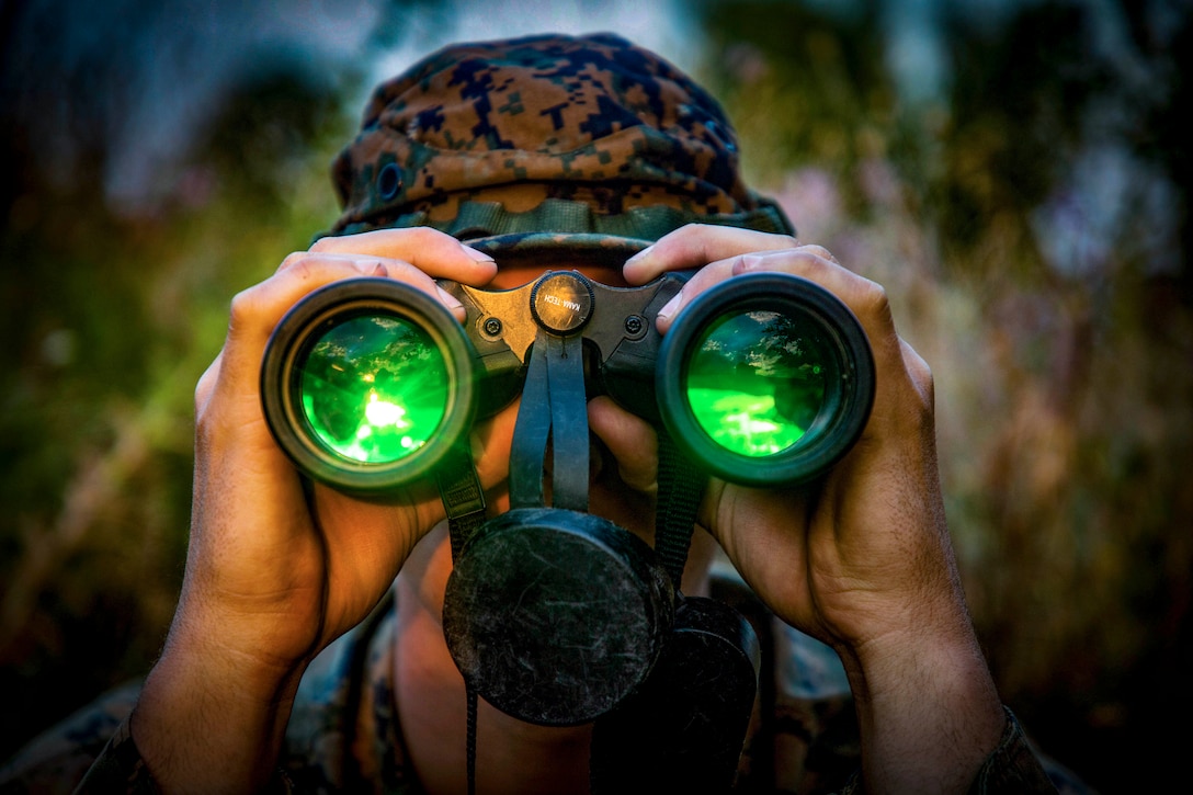 A Marine looks through binoculars with green lenses.