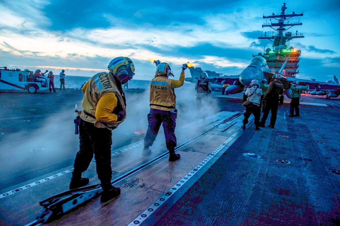 Two sailors use hand signals while standing on the deck of a ship.