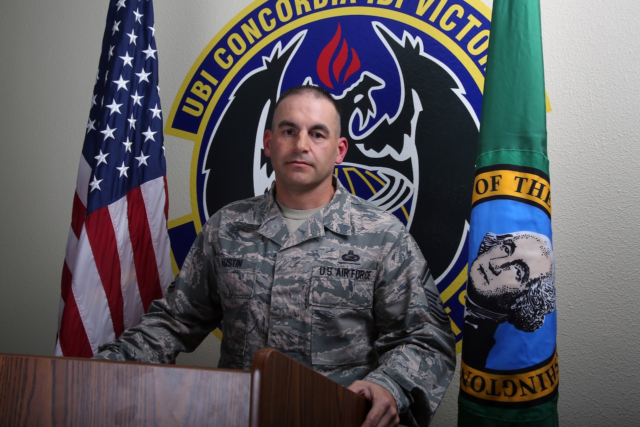 A senior enlisted airman stands behind a podium in front of flags and a unit logo.