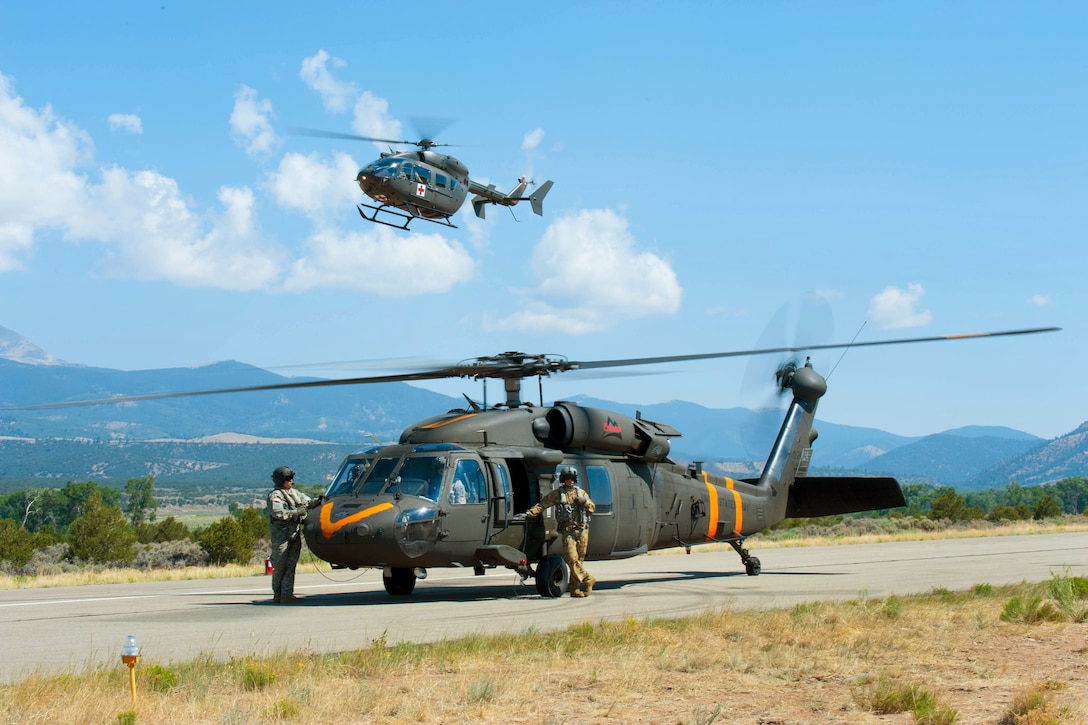 A UH-72 Lakota helicopter hovers above while soldiers prepare their UH-60 Black Hawk helicopter before departing.