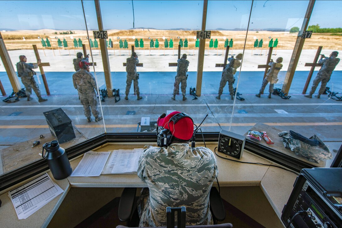 An airman behind glass watches other airmen fire weapons.
