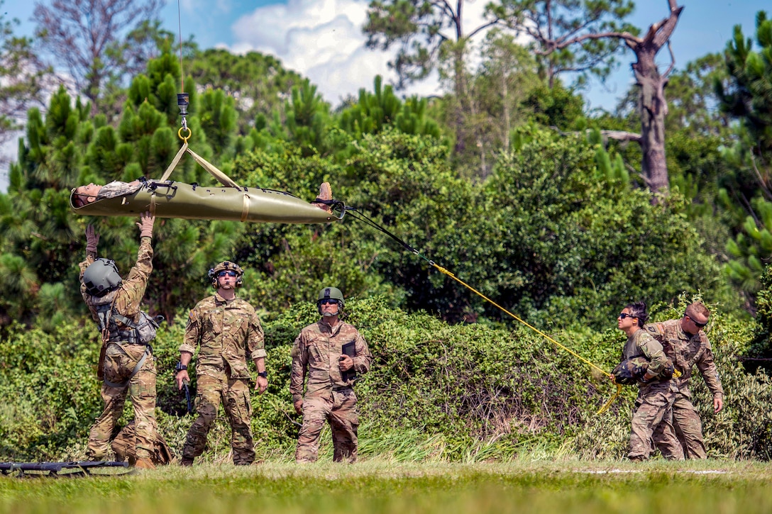 Soldiers and airmen watch as a helicopter lifts a manikin on a stretcher.