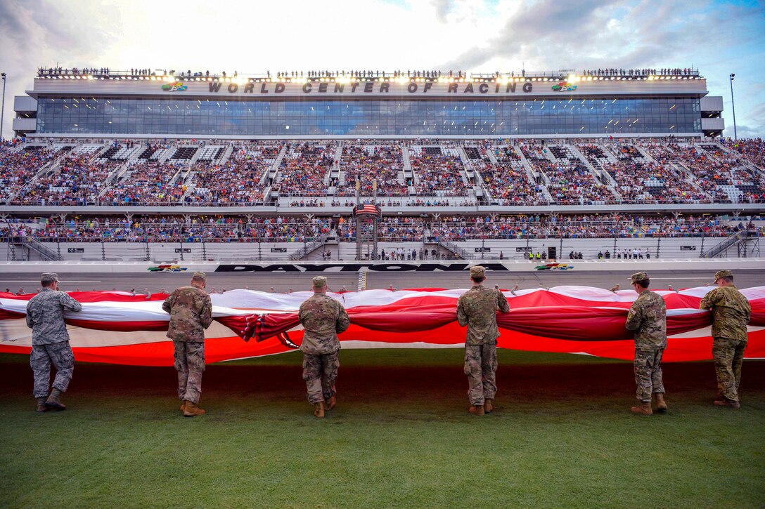 Service members gather a large American flag with a stadium in the background.