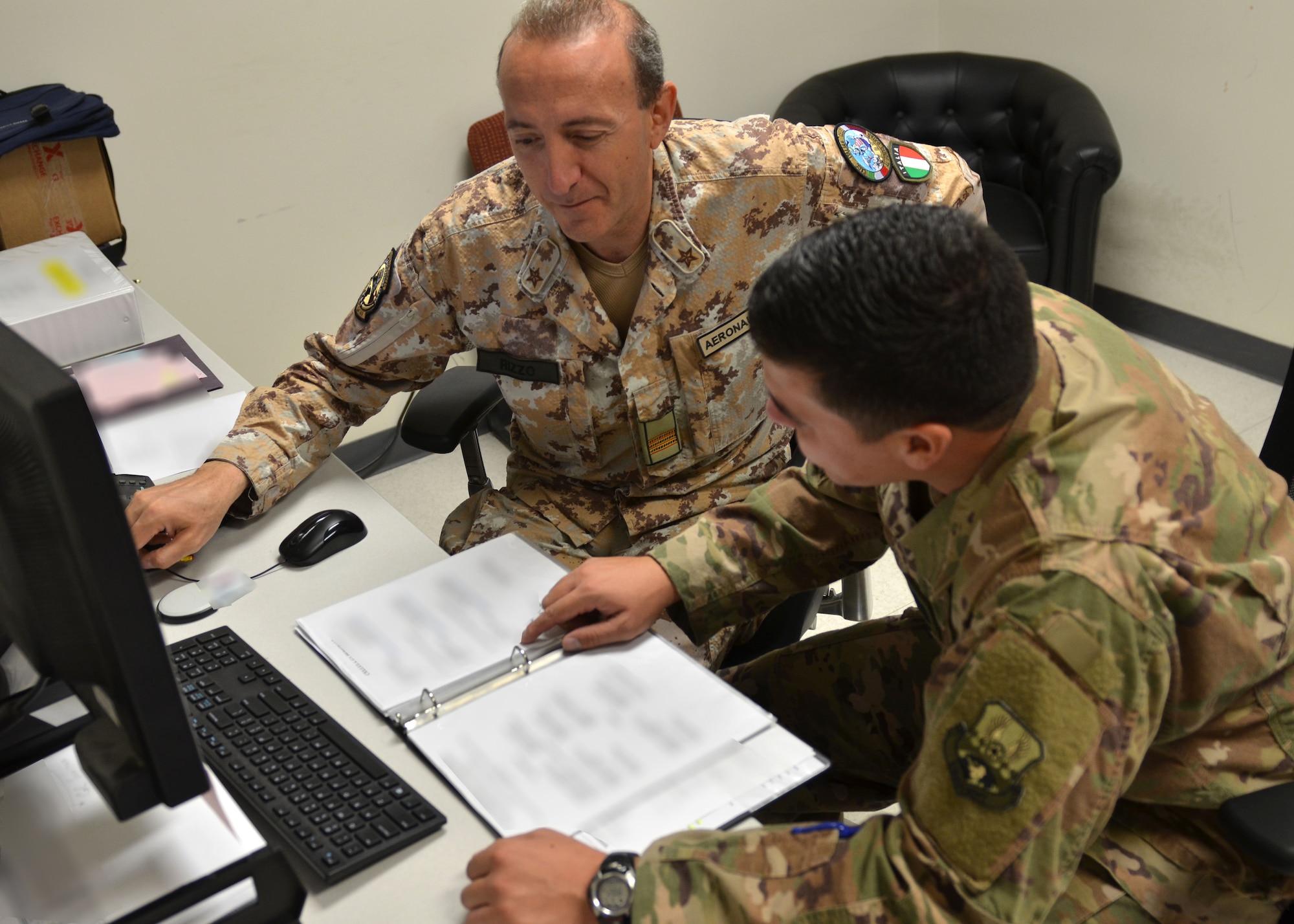 U.S. Air Force Capt. Christopher Bowser, Air Forces Central Command Coalition Coordination Cell deputy chief, looks over a document with an Italian Coalition partner, July 2, 2018 at Al Udeid Air Base, Qatar. Bowser supports more than 250 Coalition members on a daily basis.  (U.S. Air Force photo illustration by Staff Sgt. Stephanie Serrano) (This image was blurred for security reasons.)