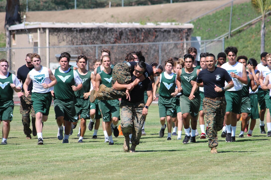 Marine Sgt. Phillip Kelso buddy carries Sgt. Kirsten Vandusen alongside Gunnery Sgt. Christopher Mondragon to demonstrate a portion of the Combat Fitness Test with students of Irvine High School, Irvine, Calif., on June 28, 2018. These football players executed the CFT with local recruiters of Marine Corps Recruiting Station Orange County to enhance their overall fitness and learn more about athleticism within the Marine Corps. (Courtesy Photo)