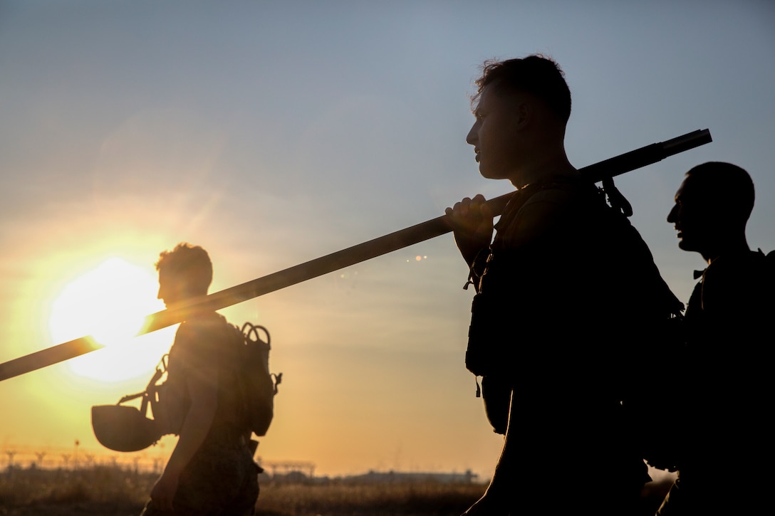 Marines are silhouetted by the sun as they carry equipment.