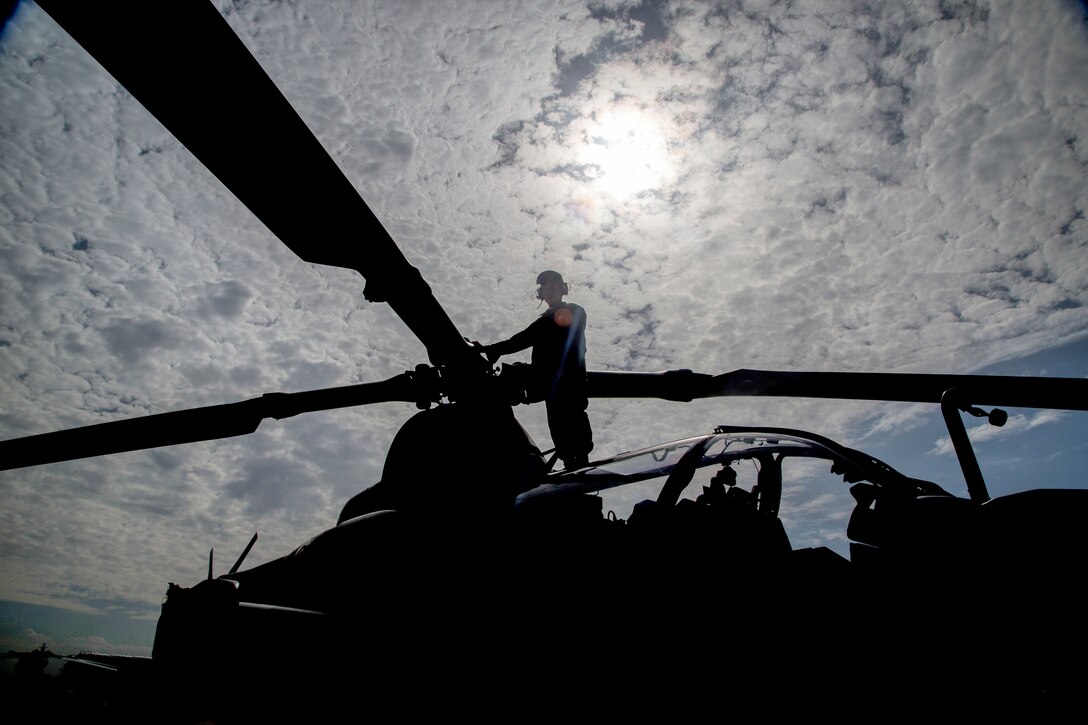 A Marine stands on top of a helicopter and is silhouetted by the sun.