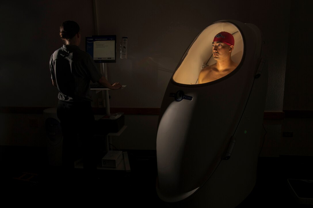 An airman stands in a lit pod-like contraption while another person looks on a screen next to him.