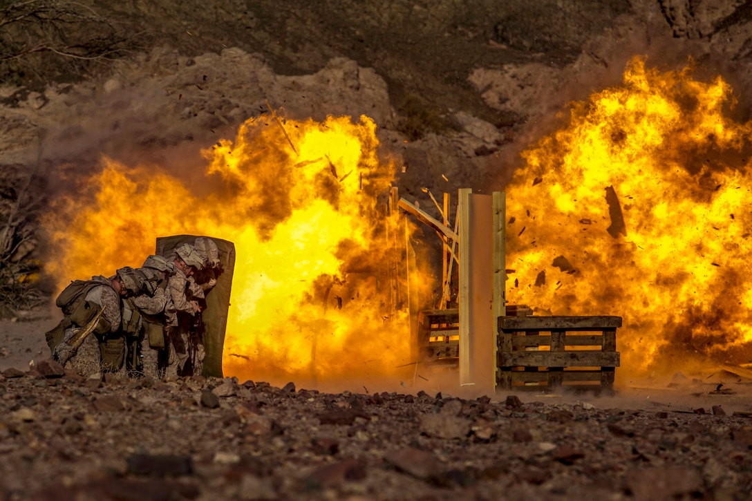 Marines tuck behind a barrier while an explosion goes off.