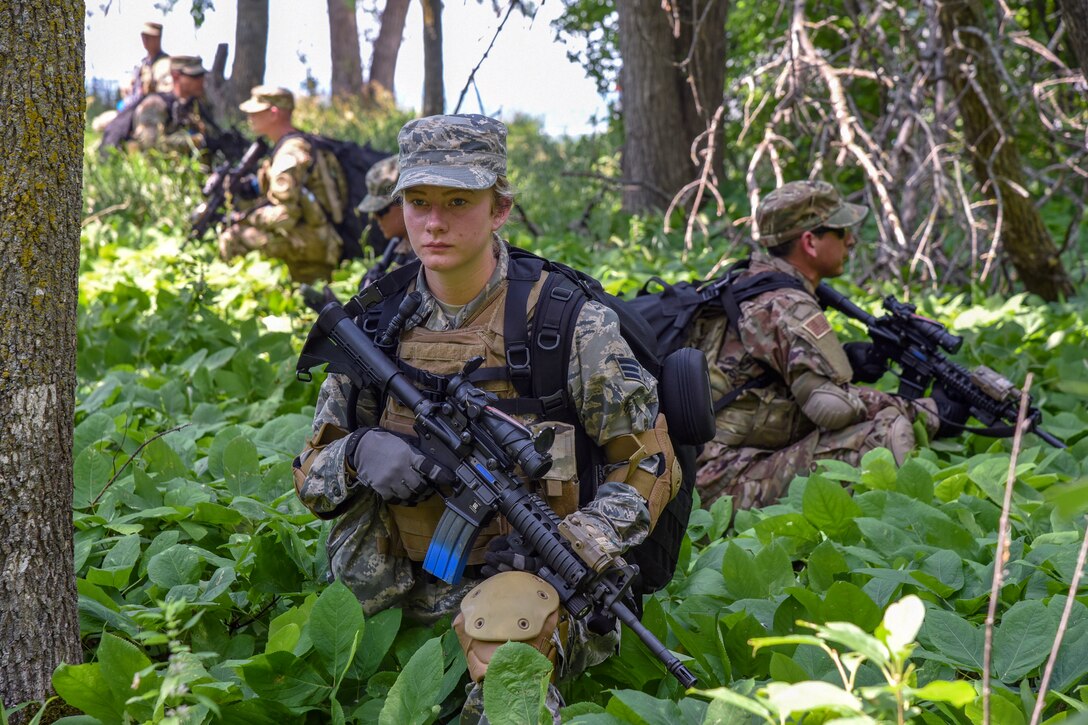 Airmen walk through a wooded area with weapons.