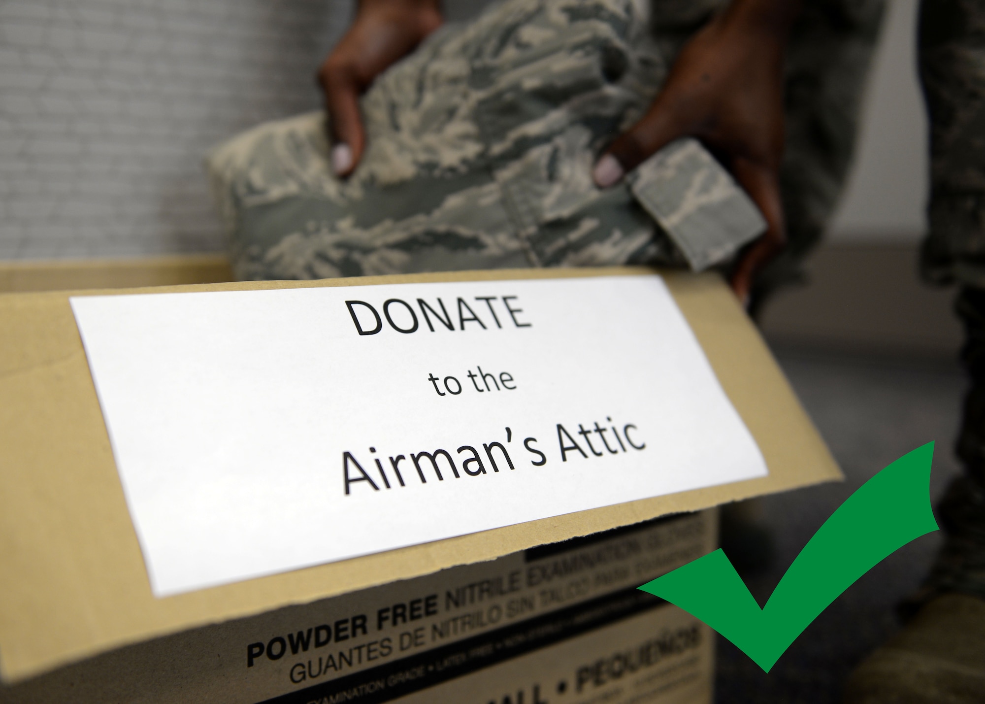 Hands are putting an Airman Battle Uniform blouse into a donation box reading "DONATE to the Airman's Attic."