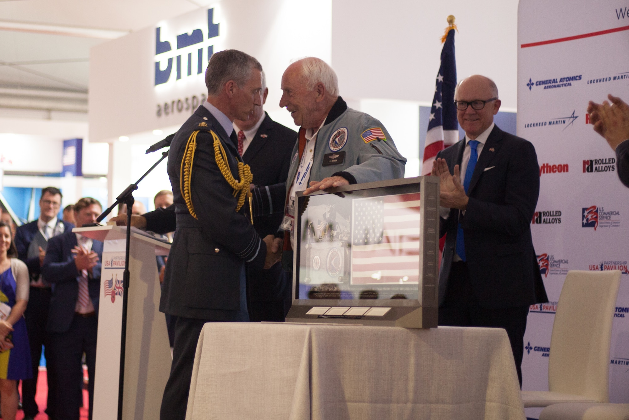 Retired Col. Al Woden, Command Module Pilot for Apollo 15, presents a flag from the 1971 lunar mission to Royal Air Force Chief of Staff, Air Chief Marshal Sir Stephen Hillier, during the Farnborough International Airshow opening ceremony July 16, 2018. By participating in the U.K-sponsored airshow, the U.S. will promote standardization and interoperability of equipment with NATO Allies and other potential coalition partners in front of more than 1,500 exhibitors from 52 countries. (U.S. Air Force photo by Capt. Allie Delury/Released)