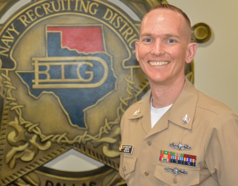 A sailor stands in front of a Navy unit logo.