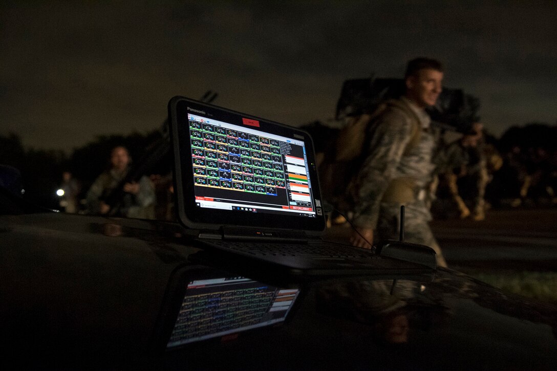 A computer screen displays data as airmen walk in the background.