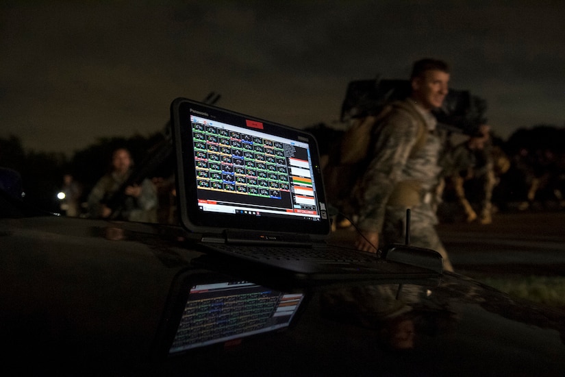 A computer screen displays data as airmen walk in the background.