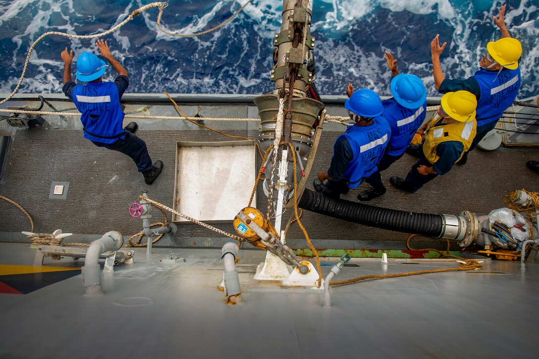 Sailors stand on the edge of a ship deck and throw a rope.