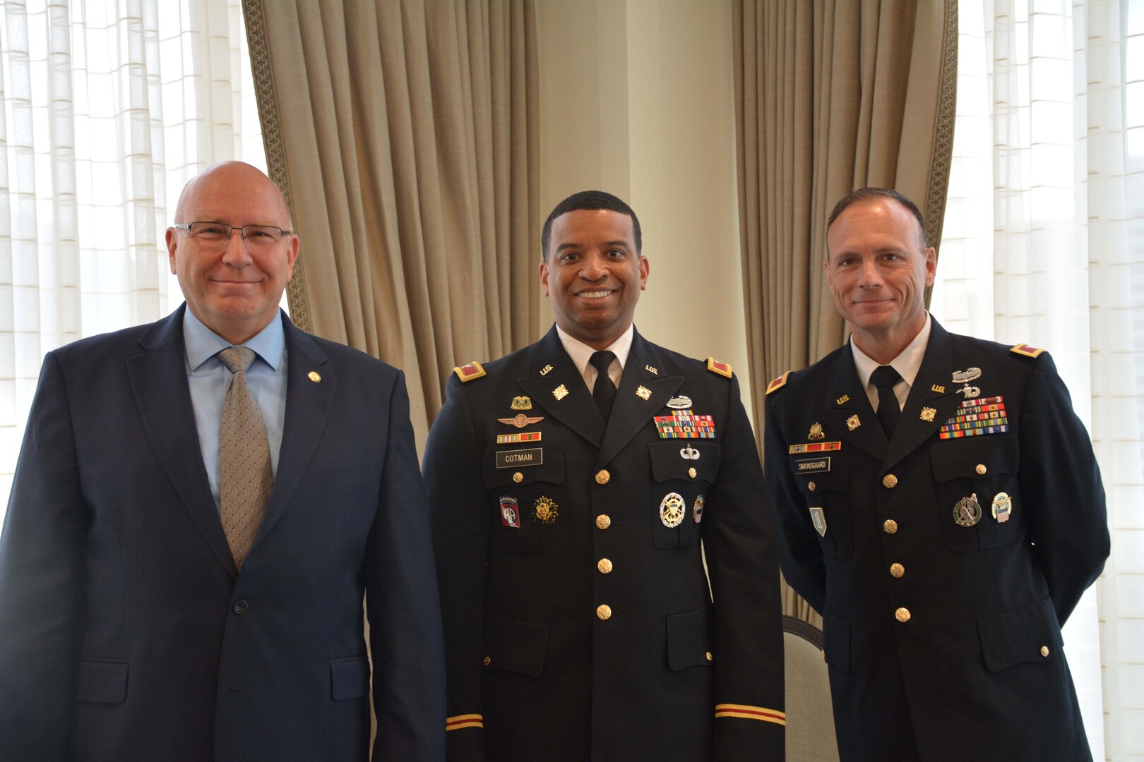 Three men stand together, one is dressed in a business suit and two are wearing Army officer dress uniforms.