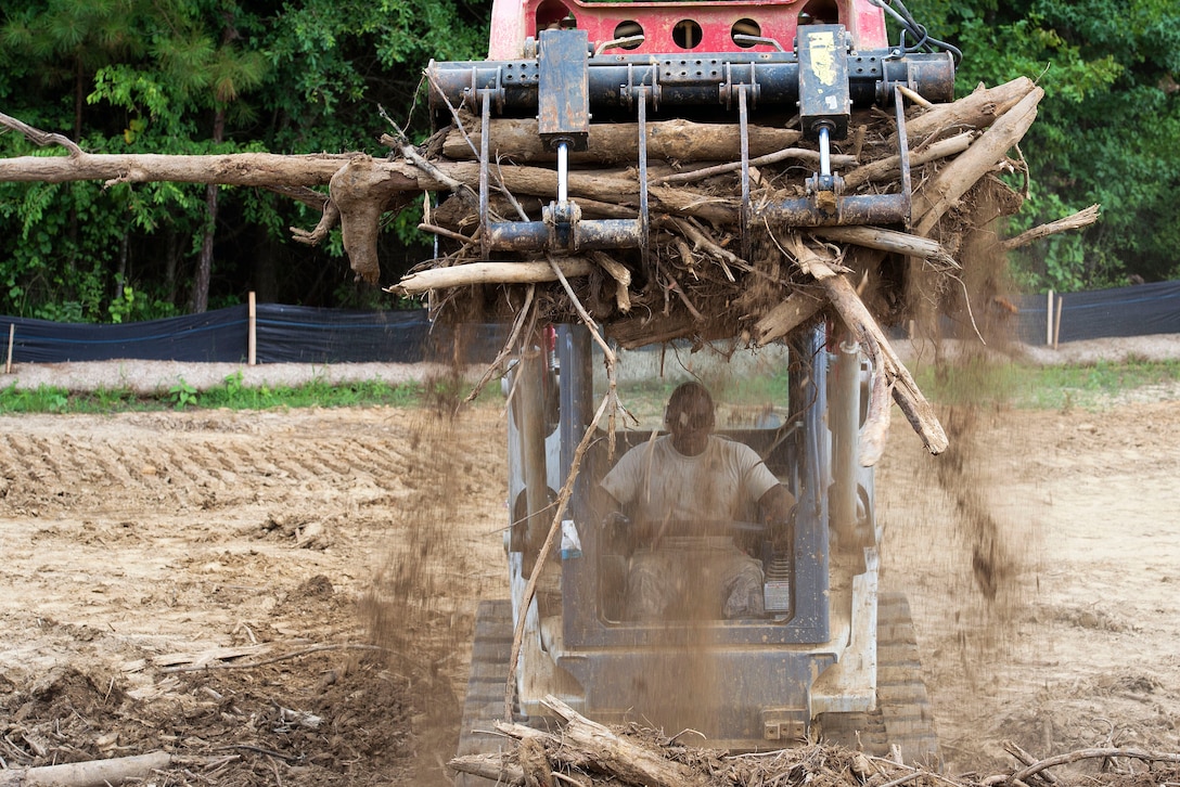 An airman shakes excess dirt off a pile wood utilizing the grappling bucket on a skid steer.