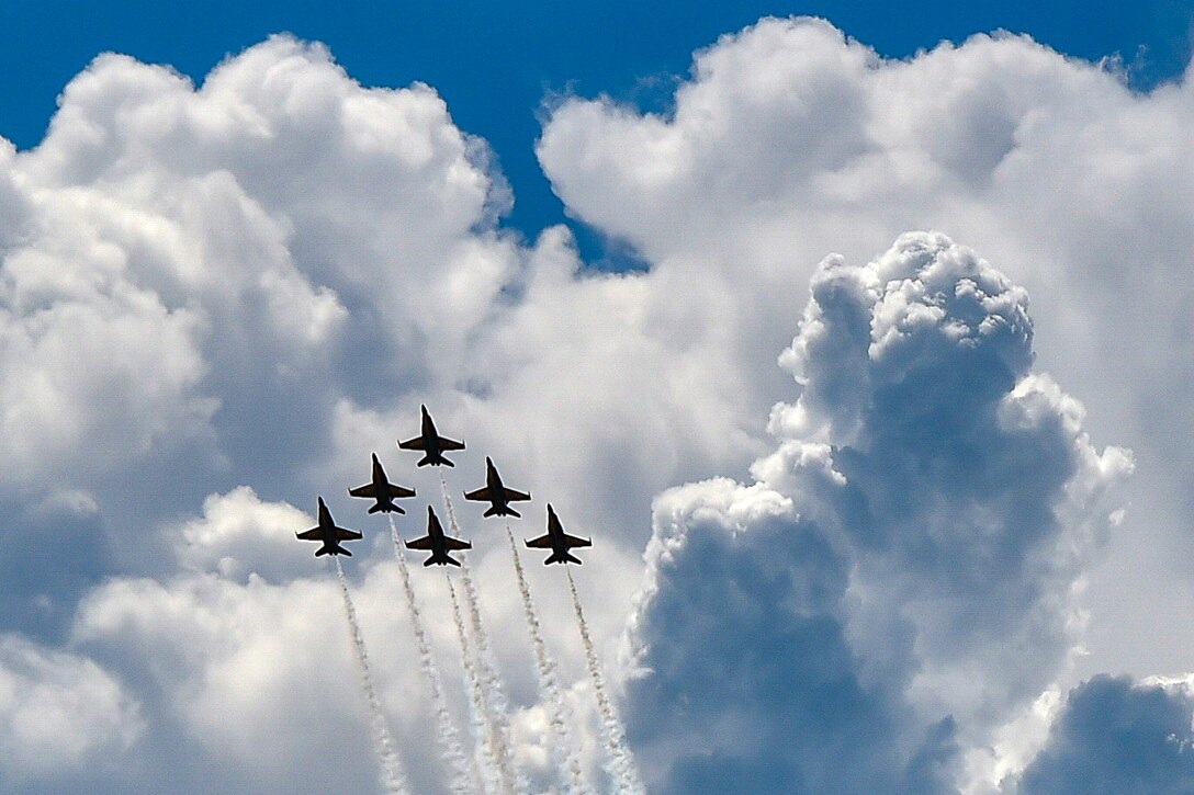 Six aircraft fly in the sky with clouds in the background.