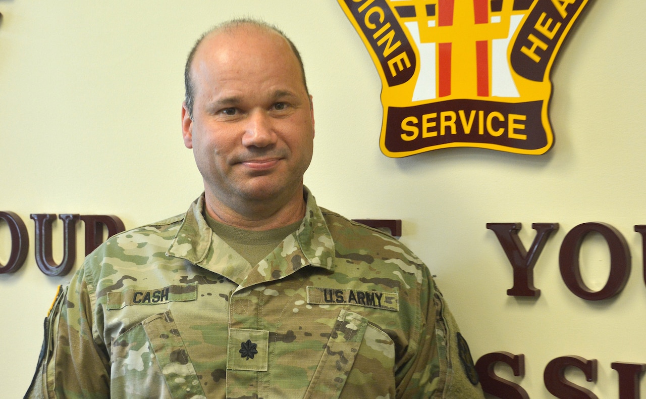 A soldier stands in front of an Army unit logo.