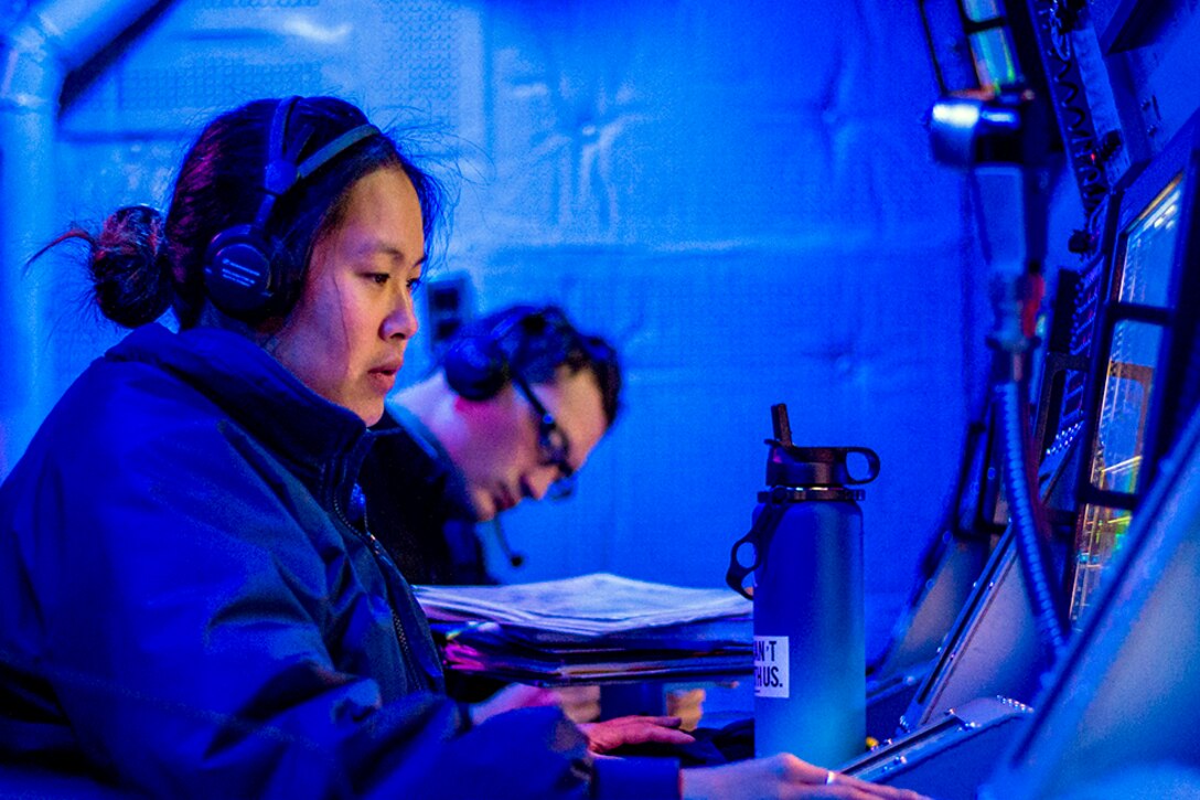 Two sailors work at screens in a blue room.
