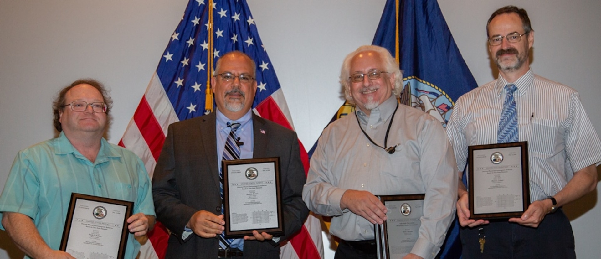 IMAGE: DAHLGREN, Va. (July 19, 2018) - John Fiore, Naval Surface Warfare Center Dahlgren Division (NSWCDD) technical director, second from left, holds a plaque reproduction of the patent certificate he presented to NSWCDD inventors - Frank Wallace, Dr. Kevin Boulais, and Blaise Corbett, left to right. They were among 32 patent holders honored before family, friends and colleagues at the NSWCDD Patent Awards ceremony. The three NSWCDD scientists along with Dr. Simin Feng and Dr. Victor Gehman were recognized for their patented innovation, 'Discrete Phased Electromagnetic Reflector Based on Two-State Elements'. This technology is an efficient means of 'steering' electromagnetic waves such as are used in radar and communications. This invention enables the warfighter to precisely guide electromagnetic beams over a broader range of frequencies than was possible before.  (U.S. Navy photo by Ryan DeShazo/Released)