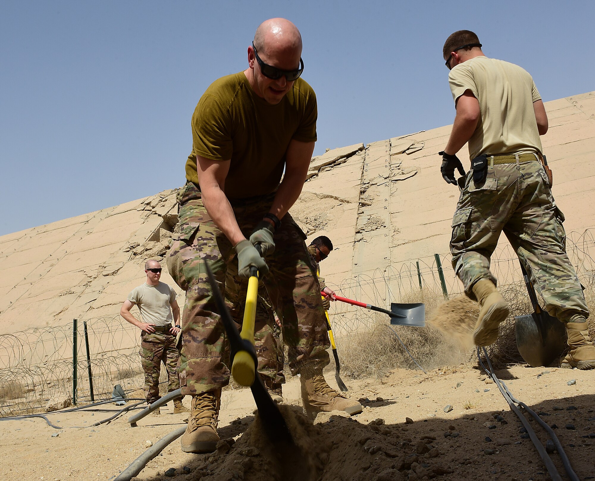 An Airman digs a hole in the sand with a pick axe