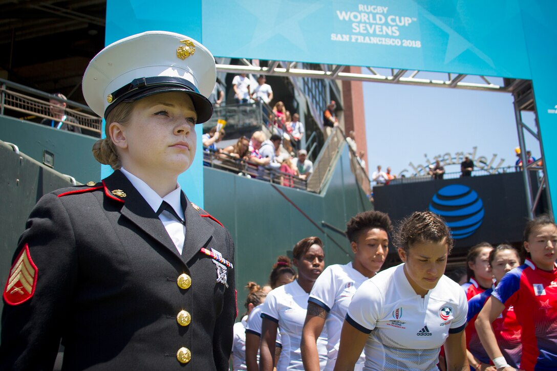 A Marines stands as rugby players run onto the field.