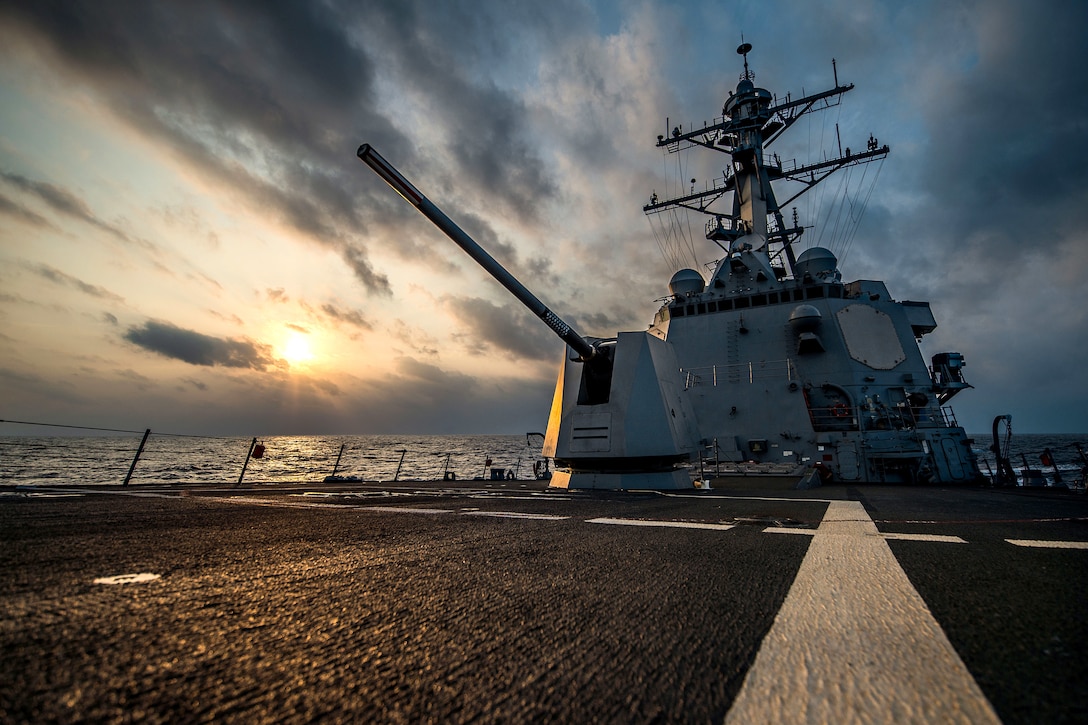 A ship sails with the sun and clouds in the background.