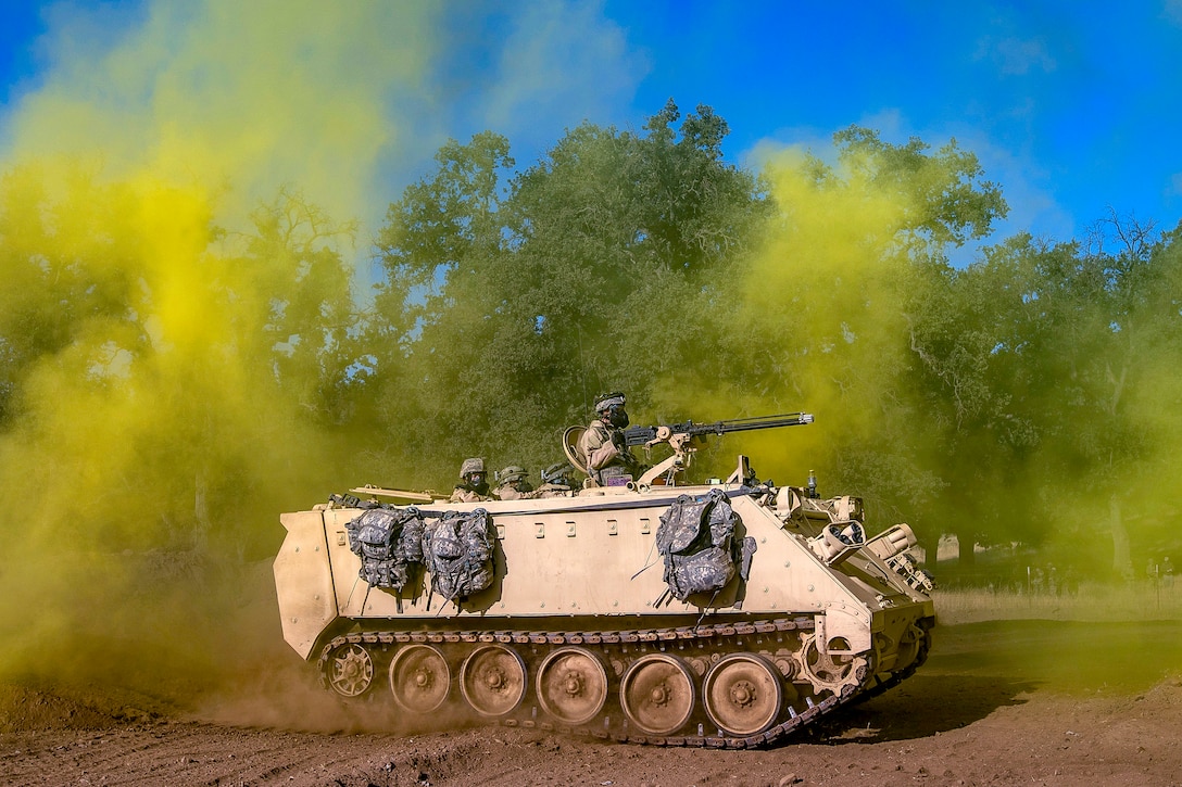 Soldiers travel in a tracked vehicle through a haze of yellow smoke.