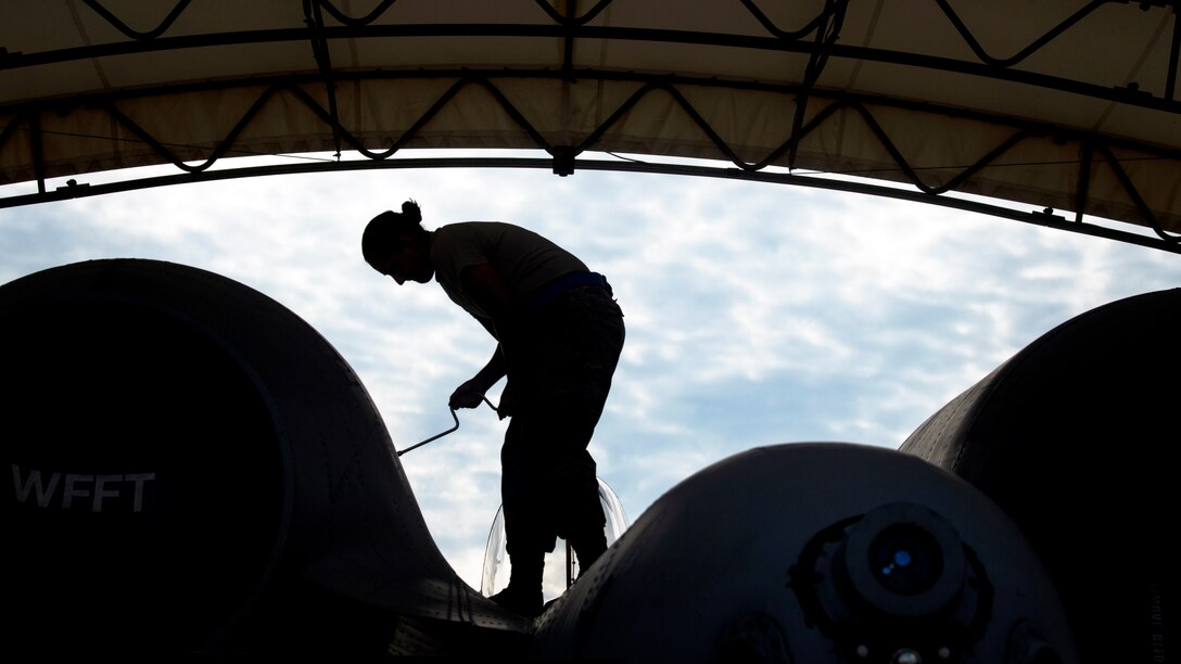 An airman, shown in silhouette, stands on an aircraft and unscrews a panel under a curved roof.
