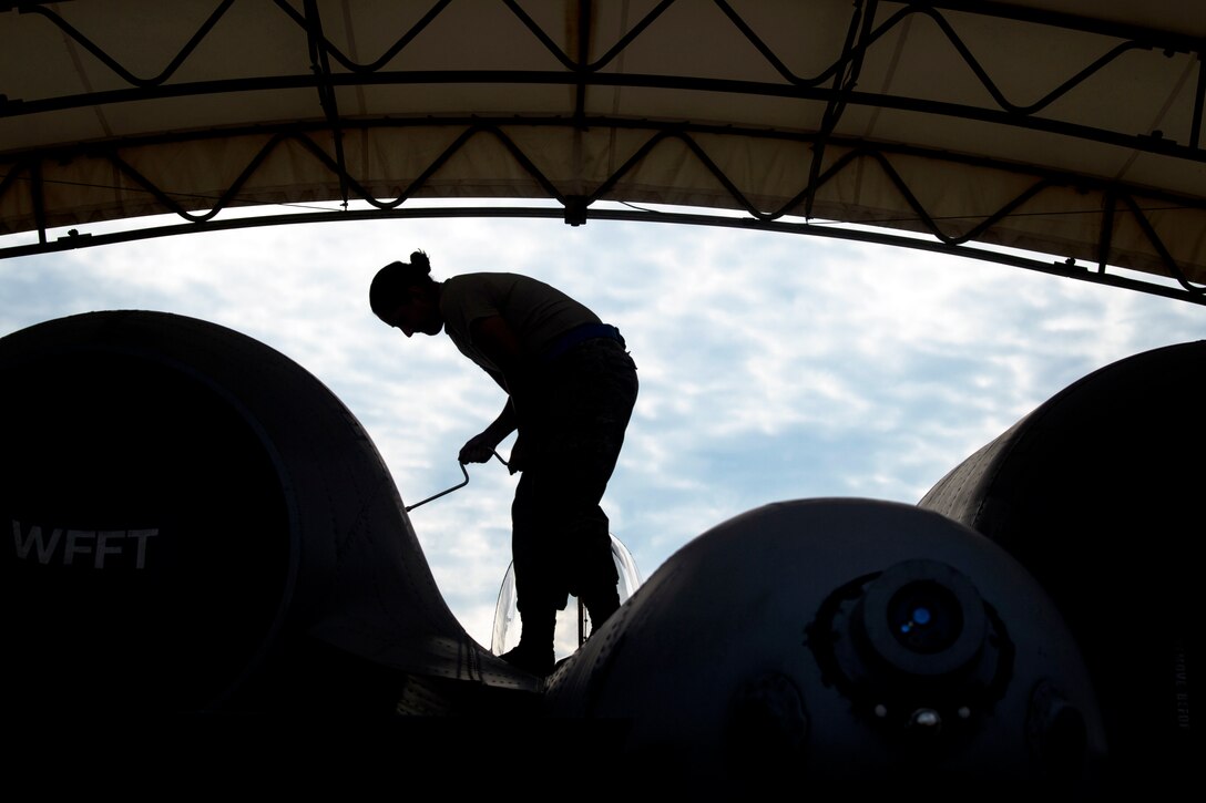 An airman, shown in silhouette, stands on an aircraft and unscrews a panel under a curved roof.