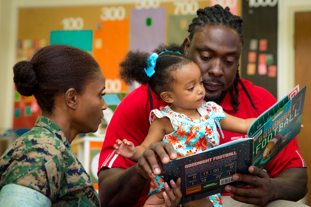 A man holds a toddler and reads her a book as a Marine watches.