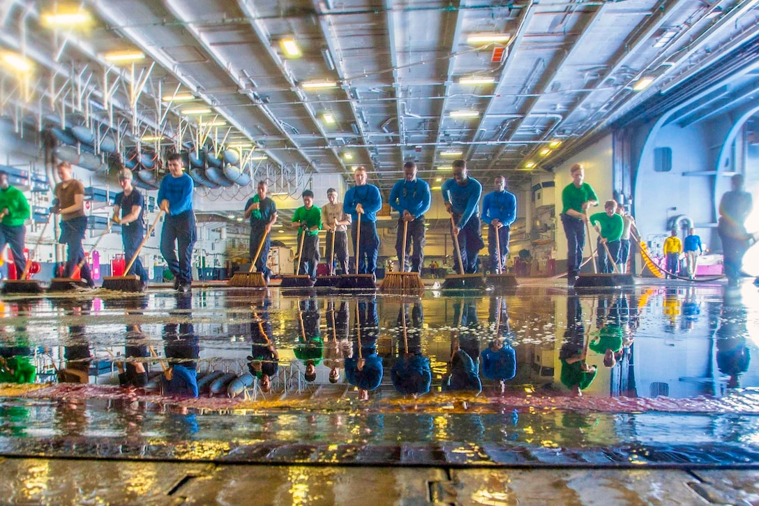A line of sailors use brooms to clean the floor.