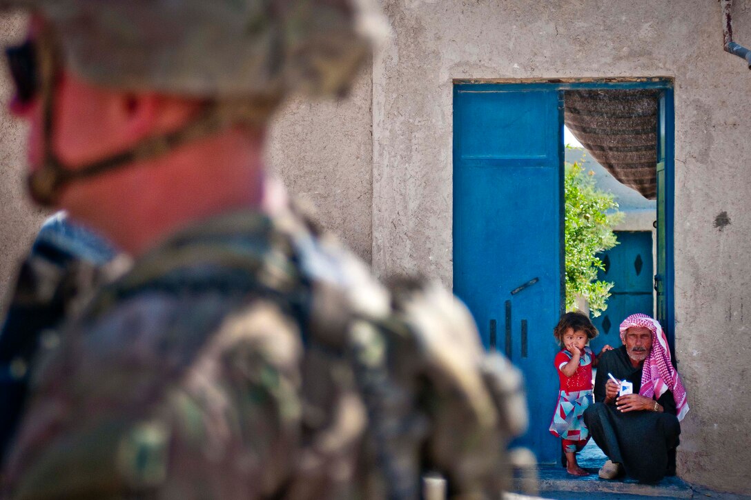 A local man and little girl watch from his house as U.S. soldiers conduct a patrol.