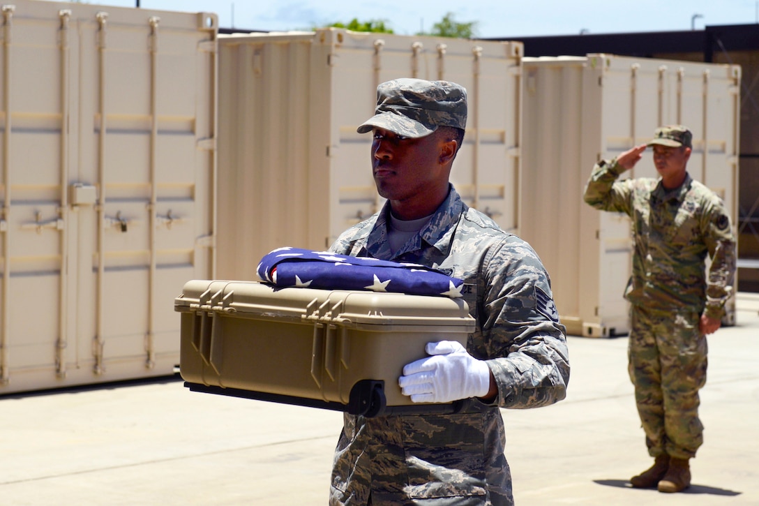 An airman carries a box of cremains with a flag folded on top, as another salutes behind him outside.