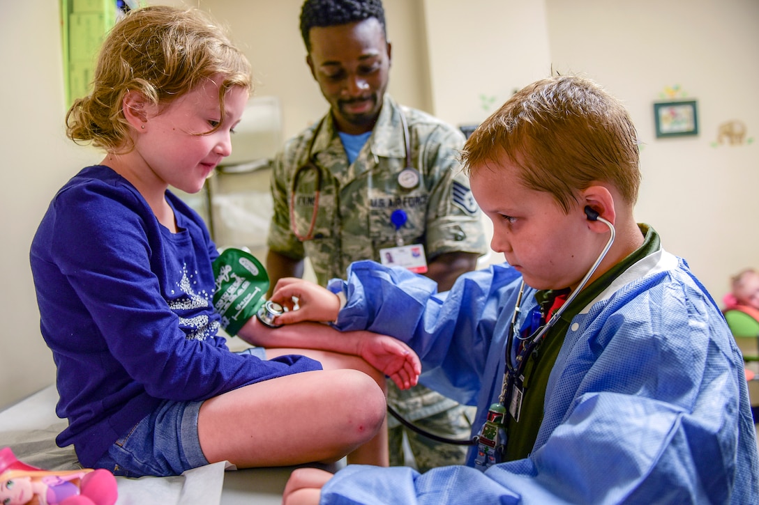 A child uses a stethoscope on the arm of another child with an airman in the background.