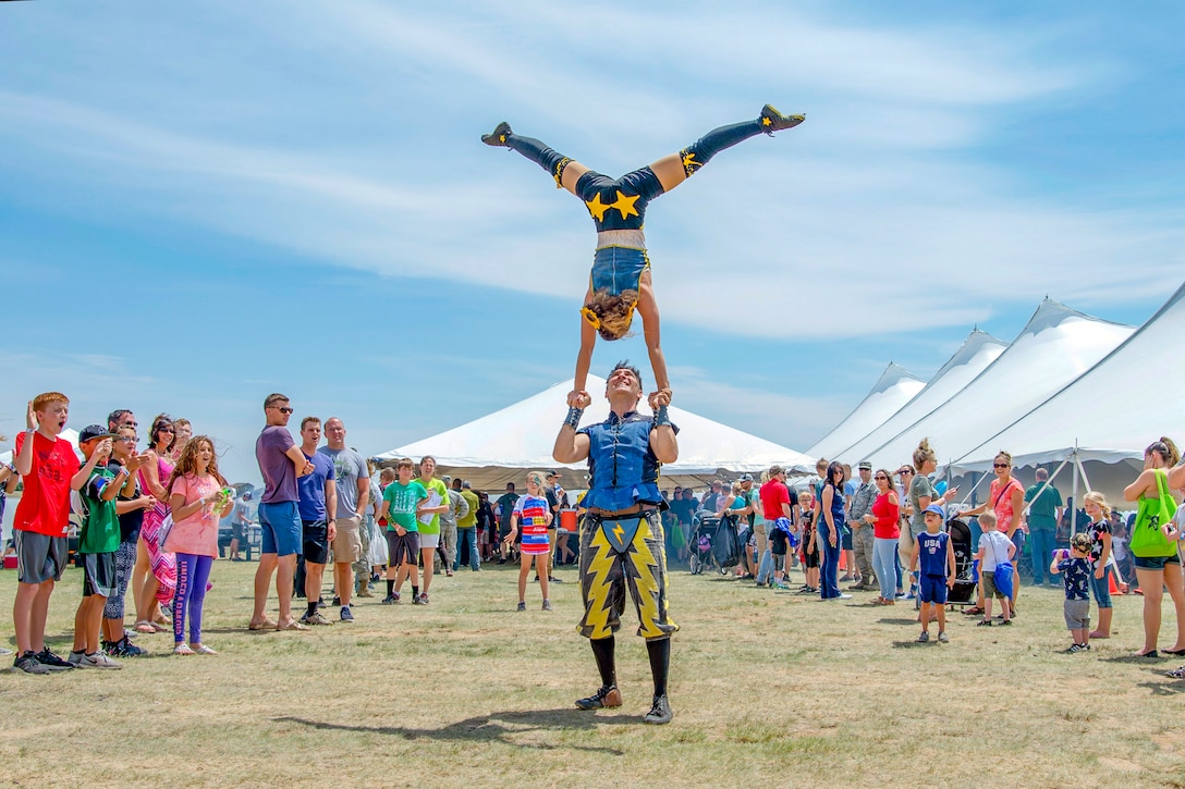 A crowd watches with visible delight as one acrobat holds another upside-down over his head in a field bordered by white tents.