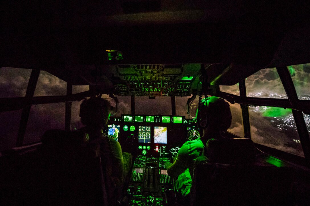 Two airmen, shown from behind, sit in a cockpit illuminated by green light.