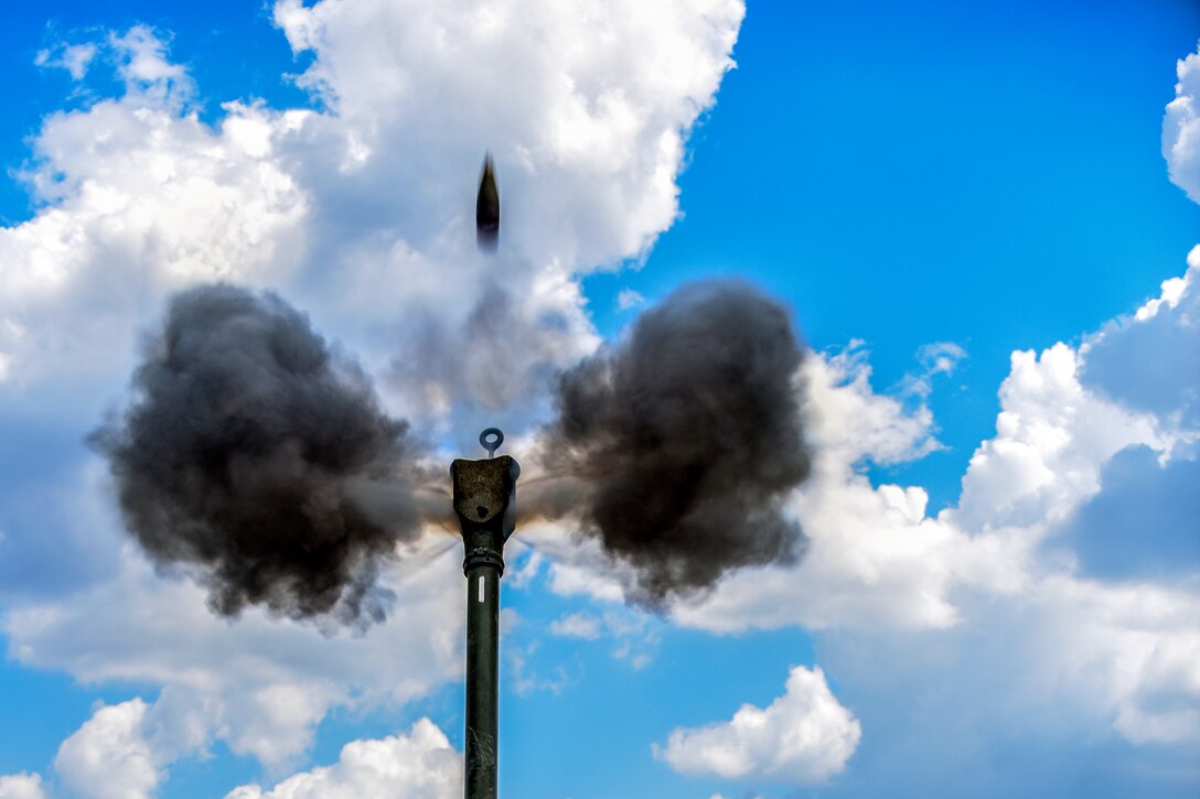 A howitzer round shoots into the blue sky as two gray puffs of smoke burst from the weapon's muzzle.