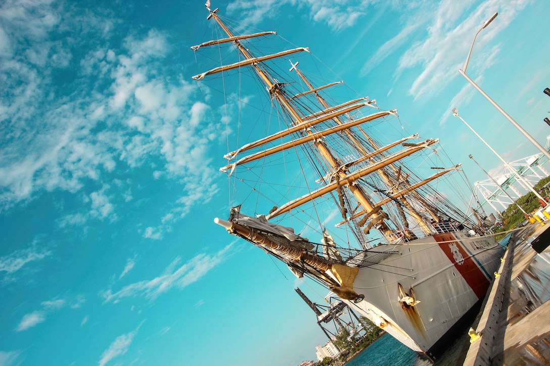 A tall ship, shown at an angle, sits moored at a pier, with bright blue sky in the background.