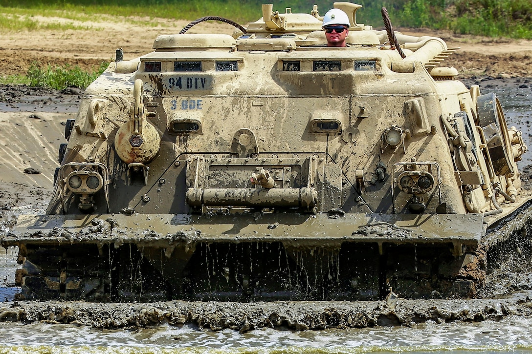 A soldier in a hard hat peers out the top of a tracked vehicle driving in mud.